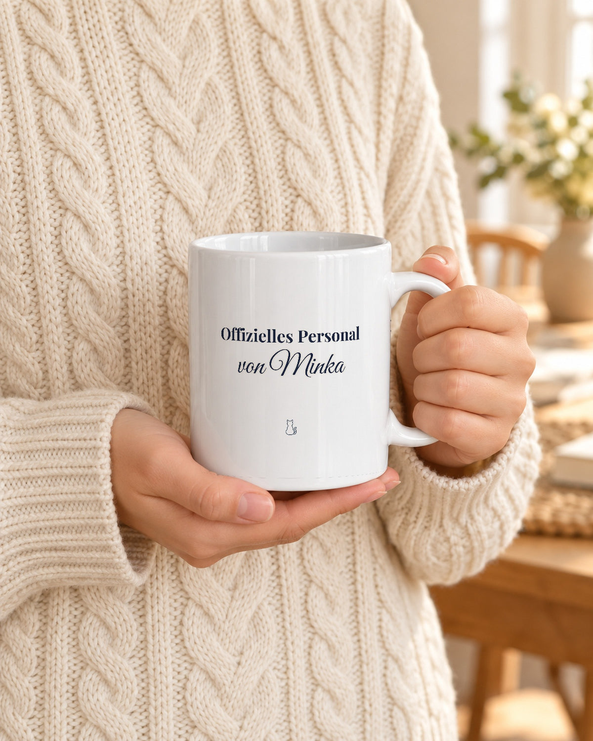 Weiße Tasse mit dem Spruch „Offizielles Personal von Minka“, gehalten von Händen im Strickpulli