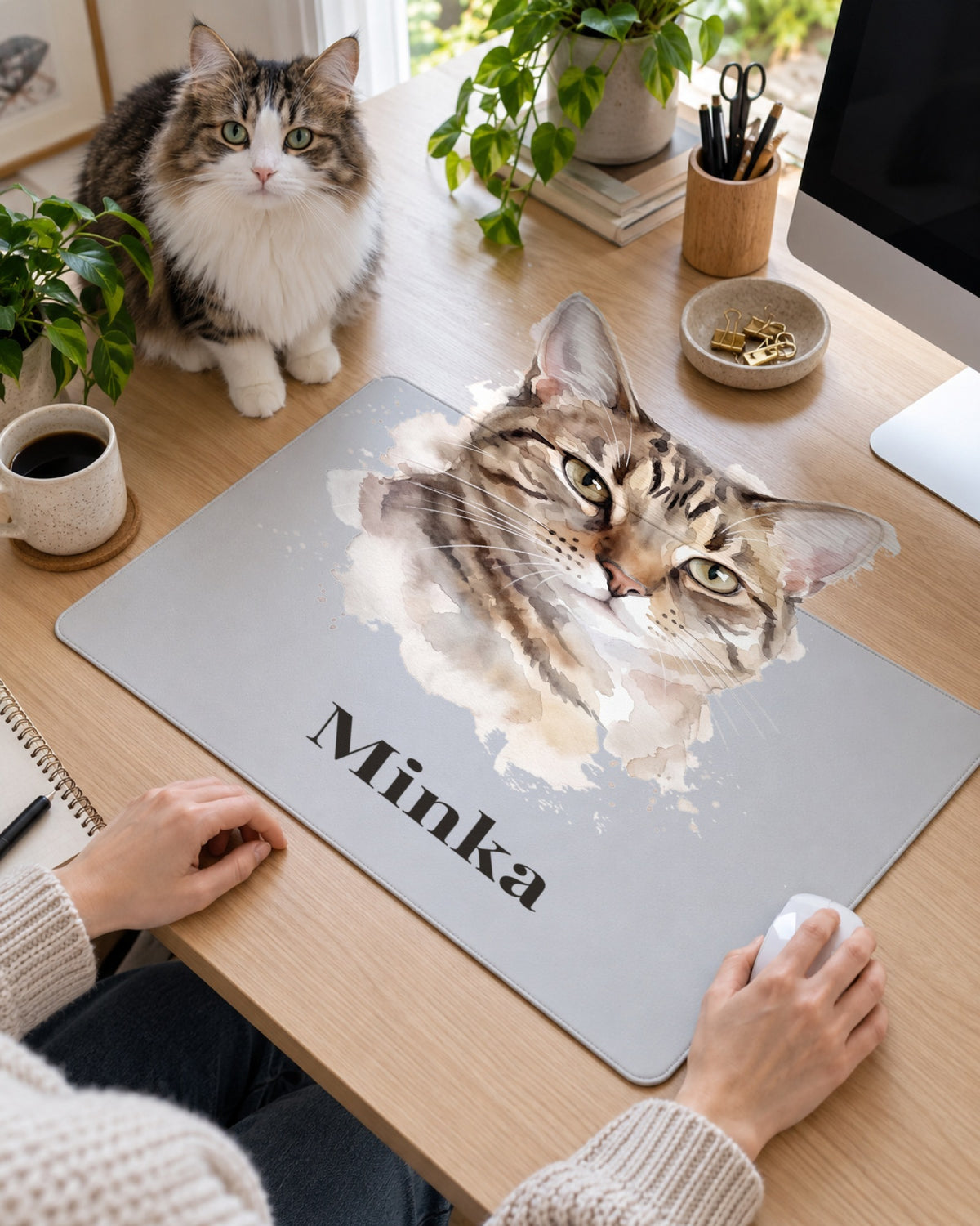 Mauspad mit Katzenportrait im Stil Aquarell und dem Namen Minka auf dem Schreibtisch, eine Katze sitzt dahinter