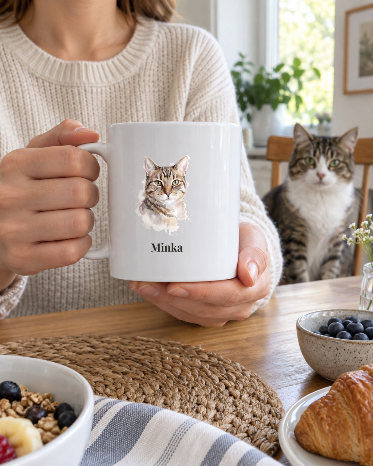 Weiße Tasse mit Tigerkatze im Aquarell-Stil und dem Namen Lotte in den Händen, die echte Tigerkatze sitzt am Frühstückstisch