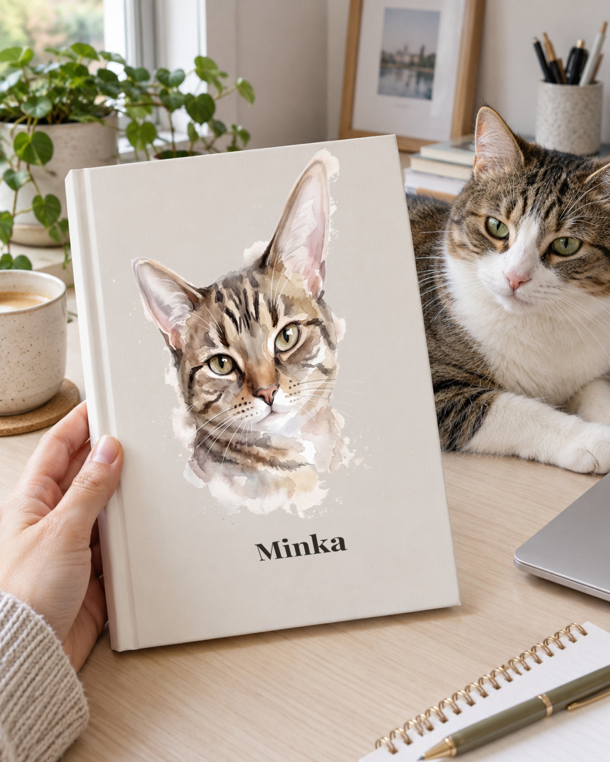 Notizbuch mit Katzenportrait im Stil Aquarell und dem Namen Minka in den Händen einer Frau, die Katze liegt auf dem Schreibtisch