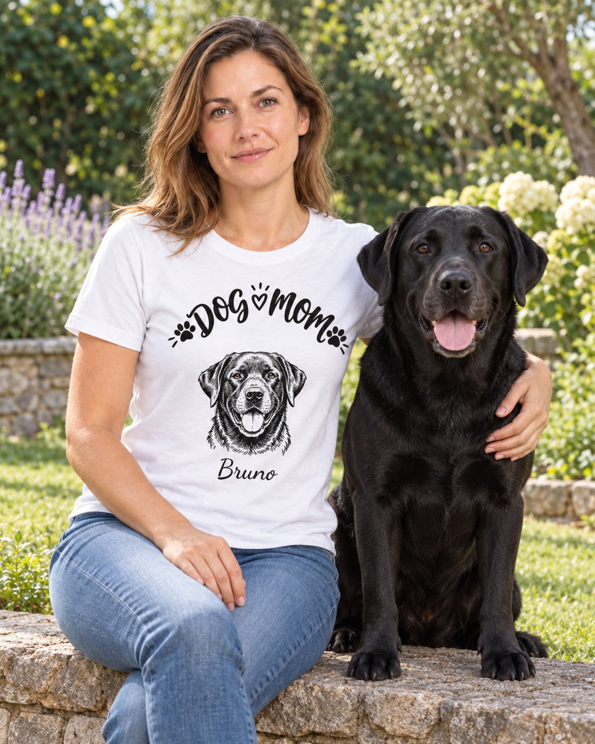 Frau trägt Dog-Mom-Shirt mit Portrait ihres schwarzen Labradors Bruno, der Hund sitzt neben ihr im Garten