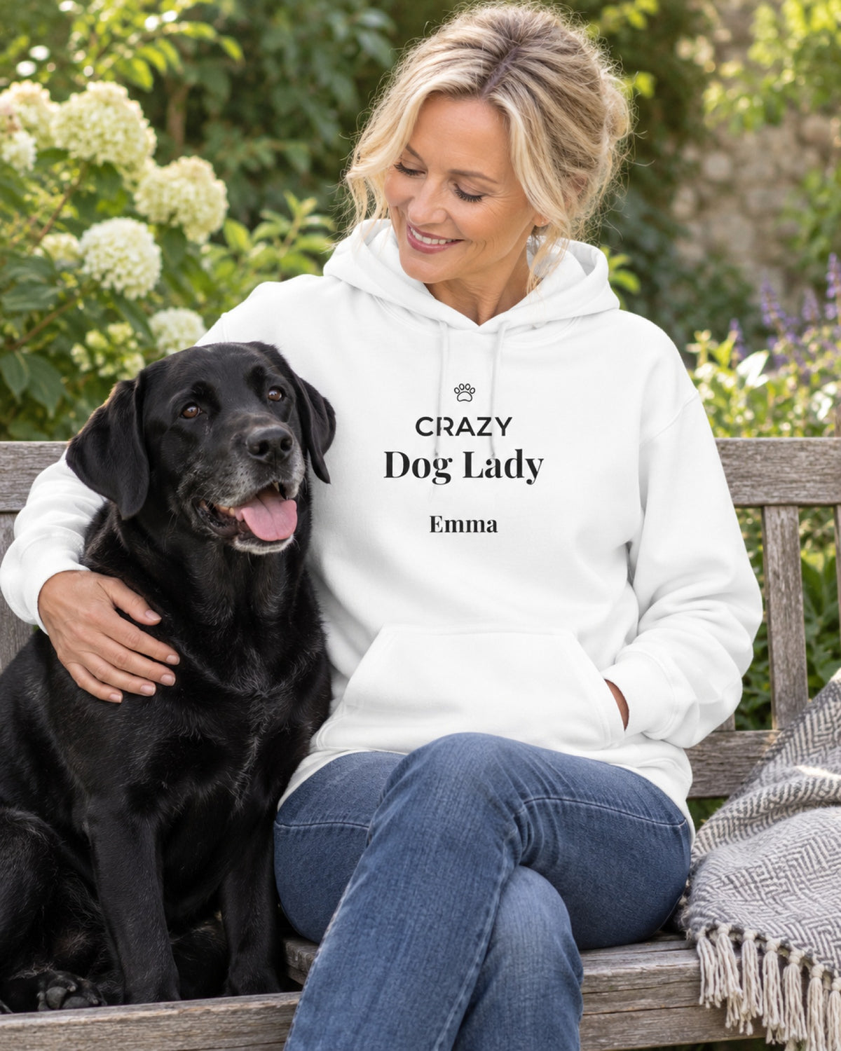 Weißer Hoodie Crazy Dog Lady mit Namen Emma, die Frau sitzt im Garten auf einer Bank mit ihrem schwarzen Labrador