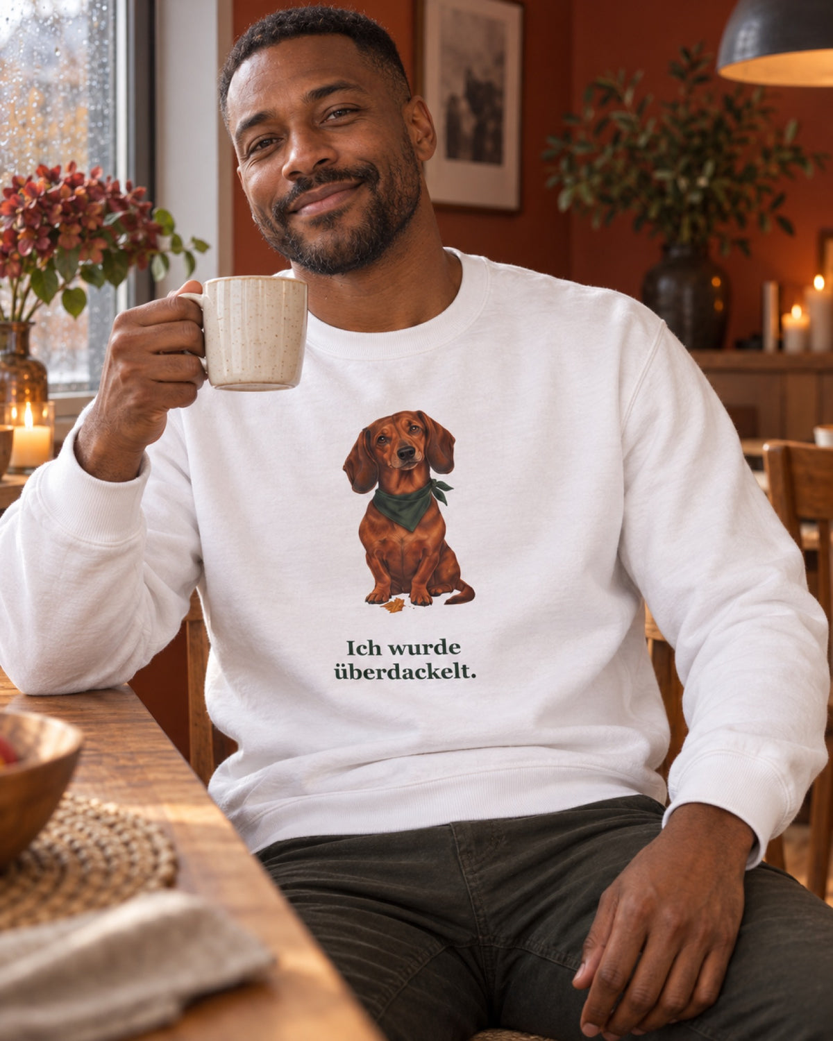 Mann im weißen Sweatshirt mit festem Dackelmotiv und dem Aufdruck Ich wurde überdackelt im rostroten Esszimmer