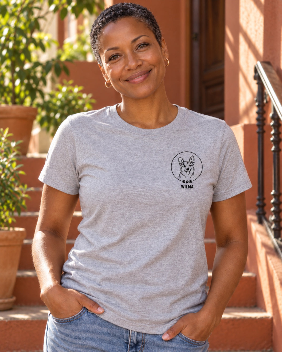Frau im graumelierten T-Shirt mit kleinem Corgi-Brustportrait und Beispielname Wilma im rostroten Treppenhaus